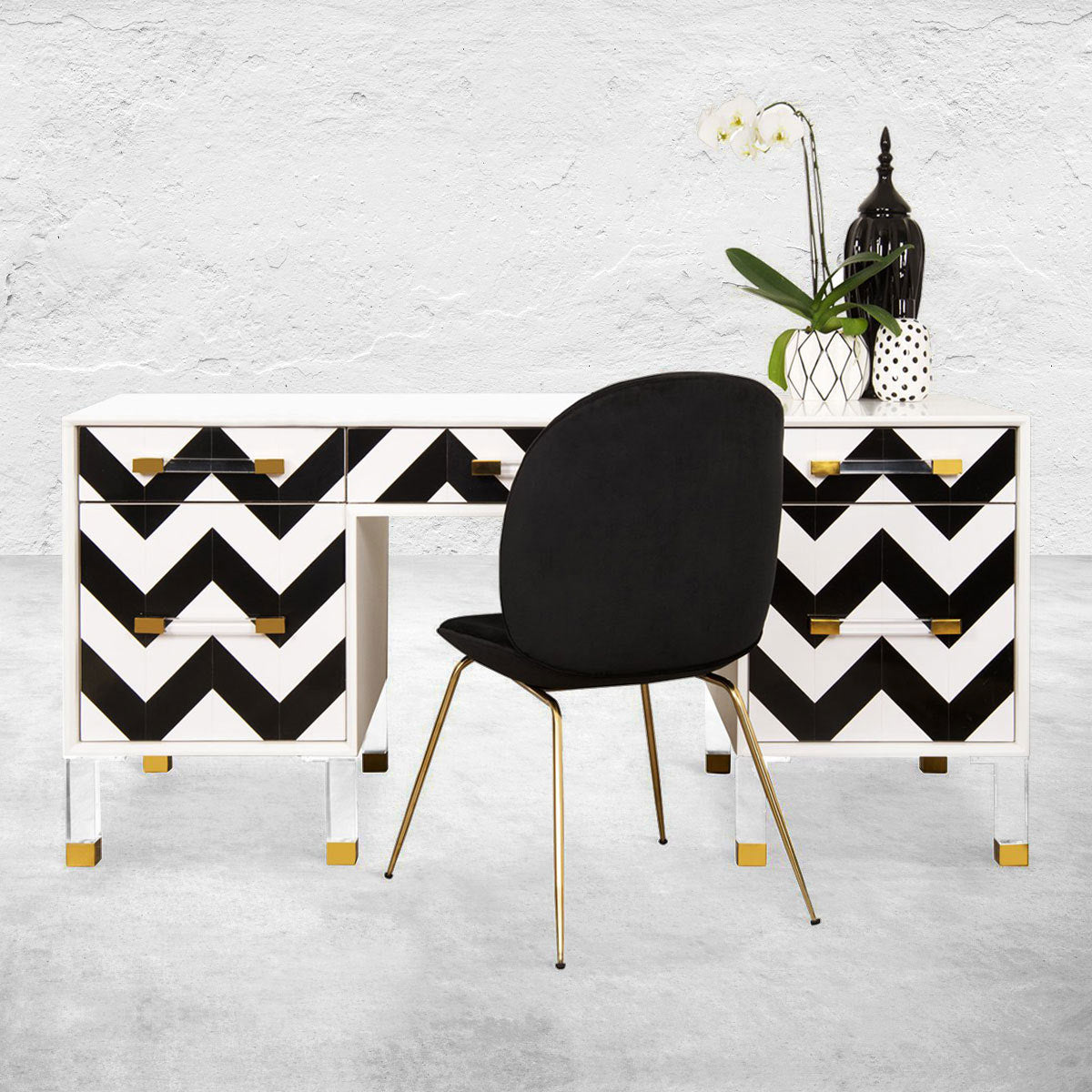 Simple black chair with metal legs in front of a six-drawer desk with a white frame, black-and-white chevron front and combination Lucite and brass legs.