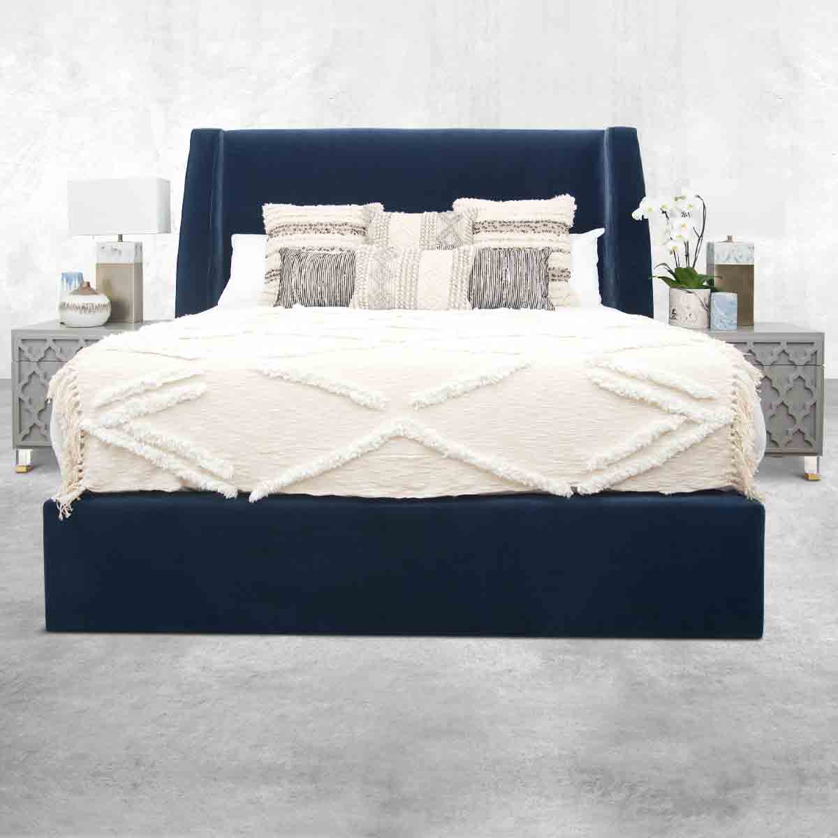 Navy upholstered platform bed with a matching headboard, white blanket, white and gray throw pillows, and gray nightstands on either side of the bed topped with vases, flowers and a lamp.